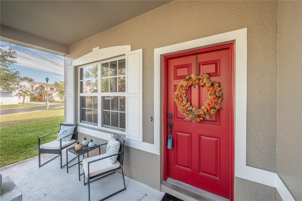 Home entrance with a red front door, autumn wreath, white shutters, and furnished covered porch.