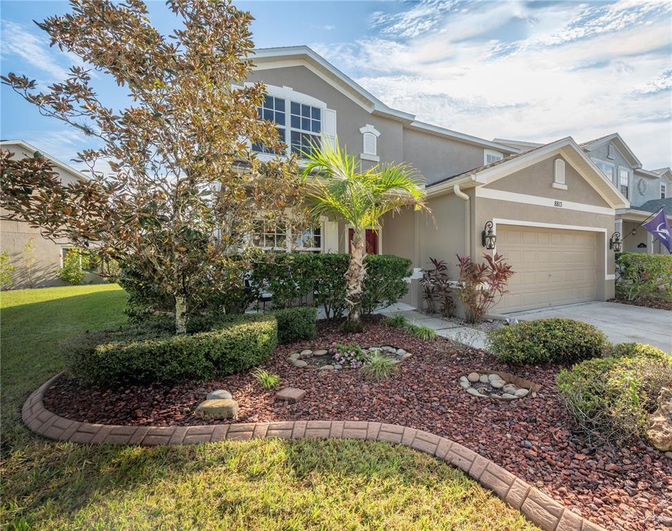 Two-story beige home with a double garage, palm tree, and landscaped front garden.