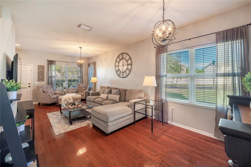 Living room with a gray sectional, patterned armchairs, hardwood floors, and large curtained windows.