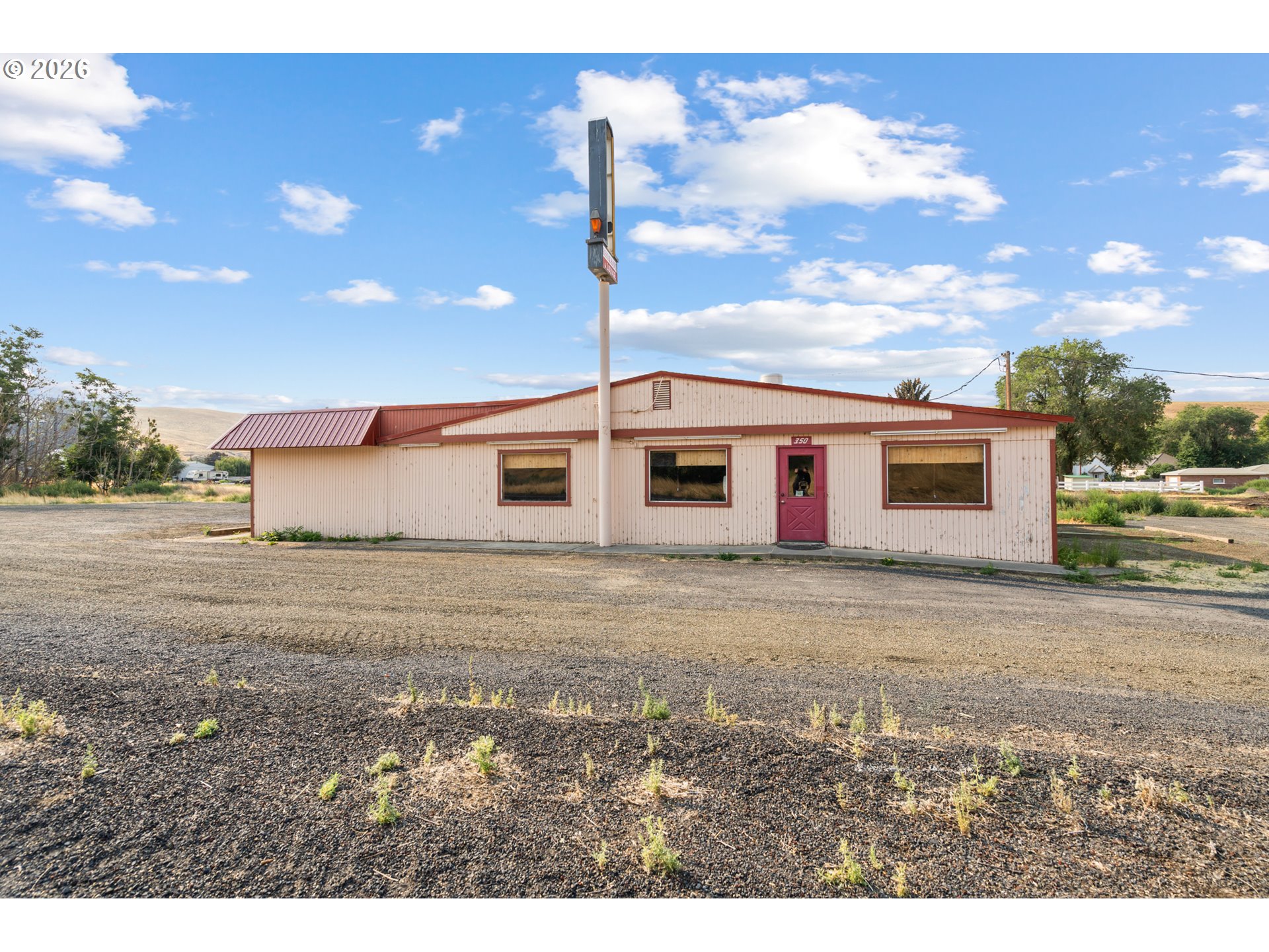 Single-story commercial building with a red metal roof and entry door beside a broad gravel lot.