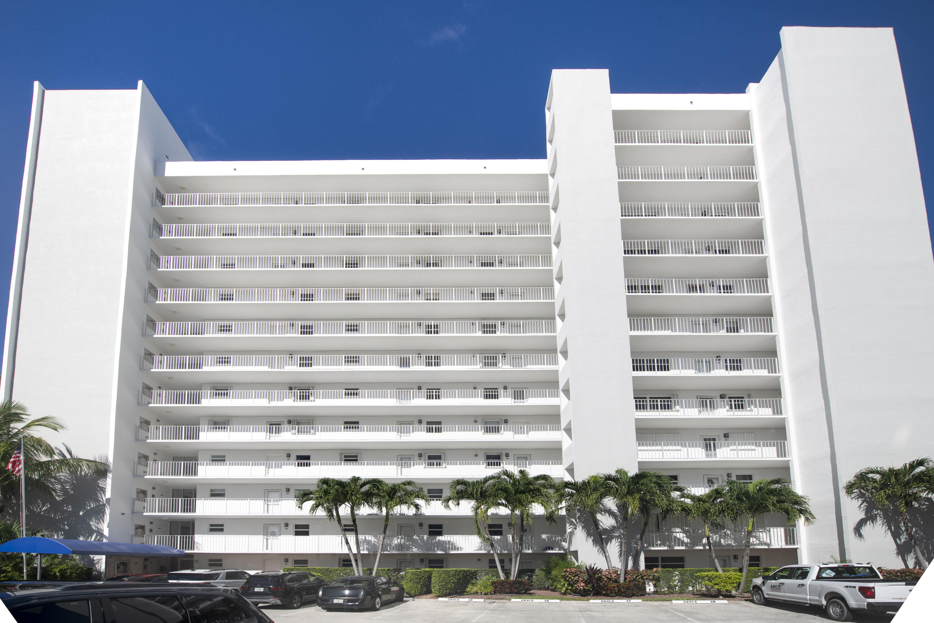 White high-rise condominium with stacked balconies, palm trees, and parking in front.