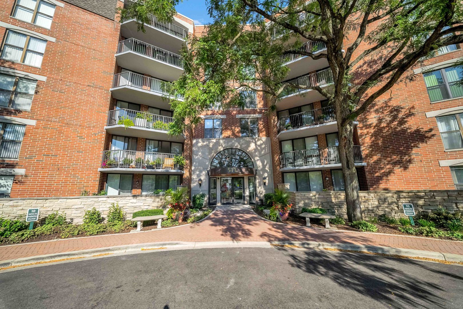 Red-brick condominium building with balconies and an arched entrance marked 511, shaded by mature trees.