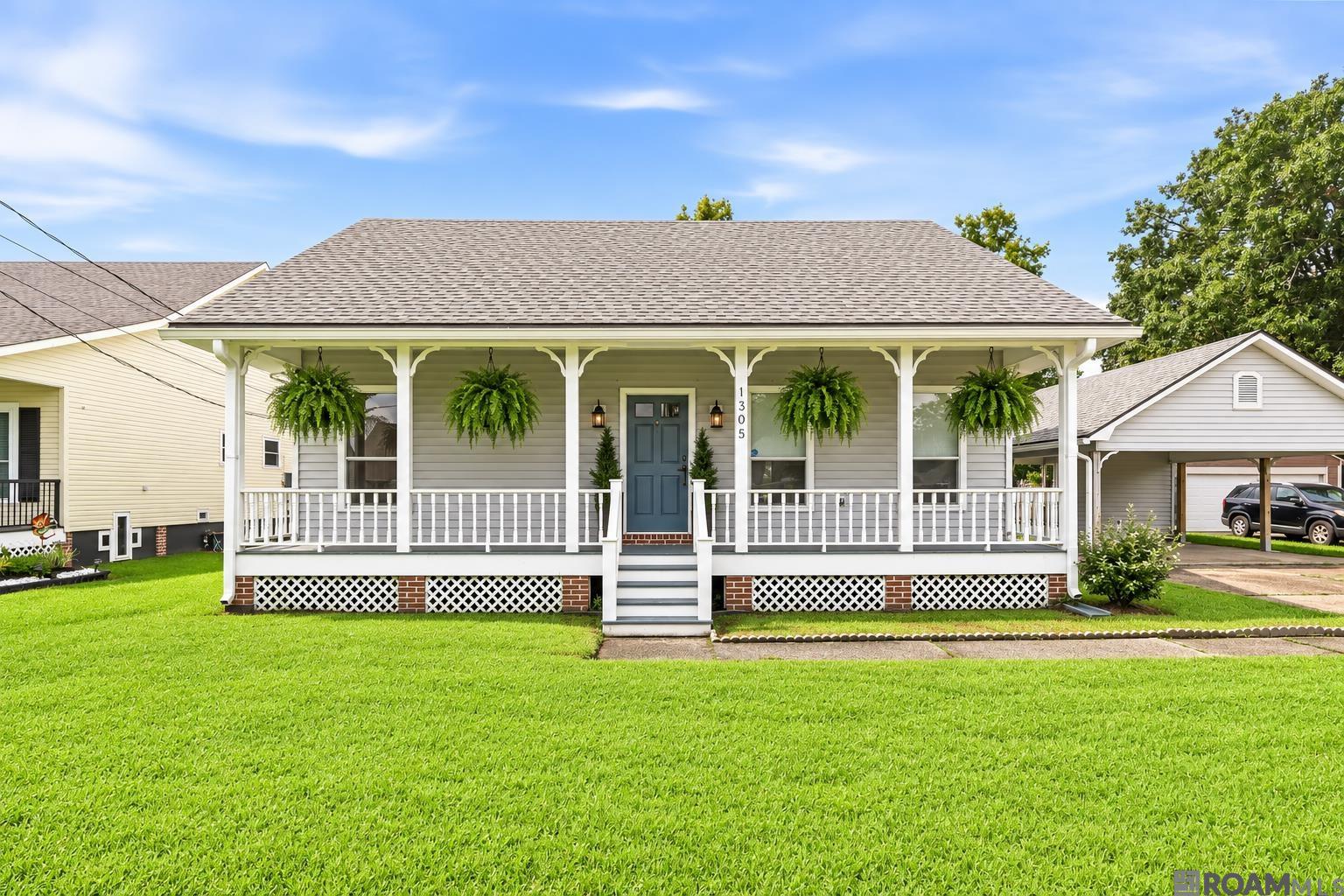Light-gray raised house with a broad porch, hanging ferns, blue front door, and green lawn.