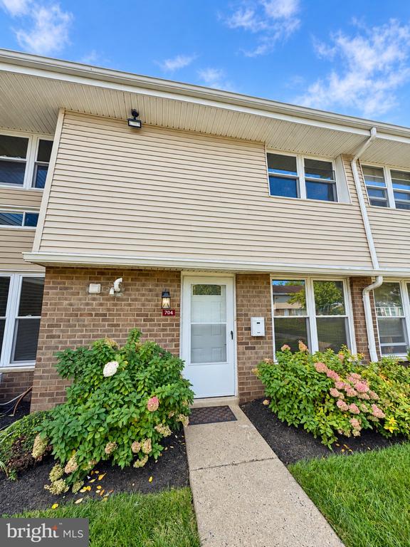 Two-story townhouse with a white entry door, concrete walkway, and flowering shrubs.