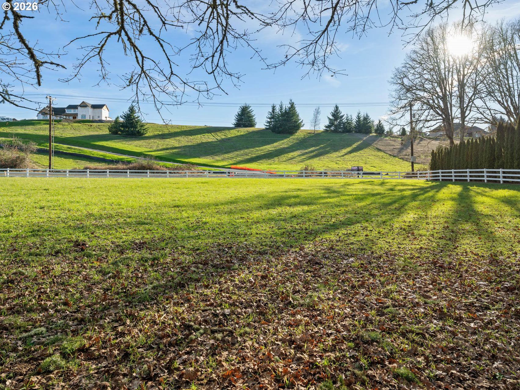 Grassy hillside lot behind a white fence, with a house on the ridge and long tree shadows.