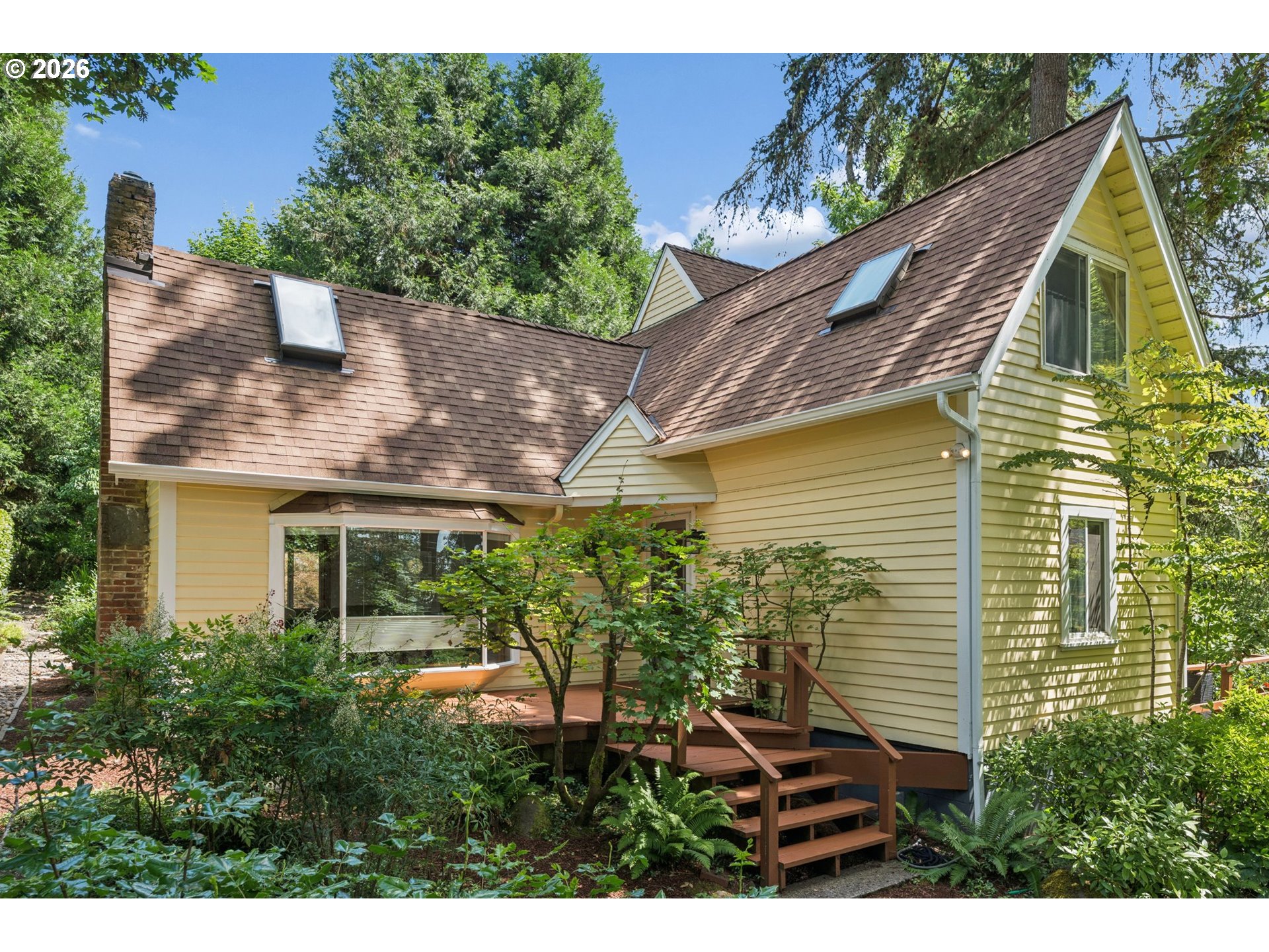 Pale yellow house with a steep shingle roof, skylights, and a raised wooden deck amid trees.