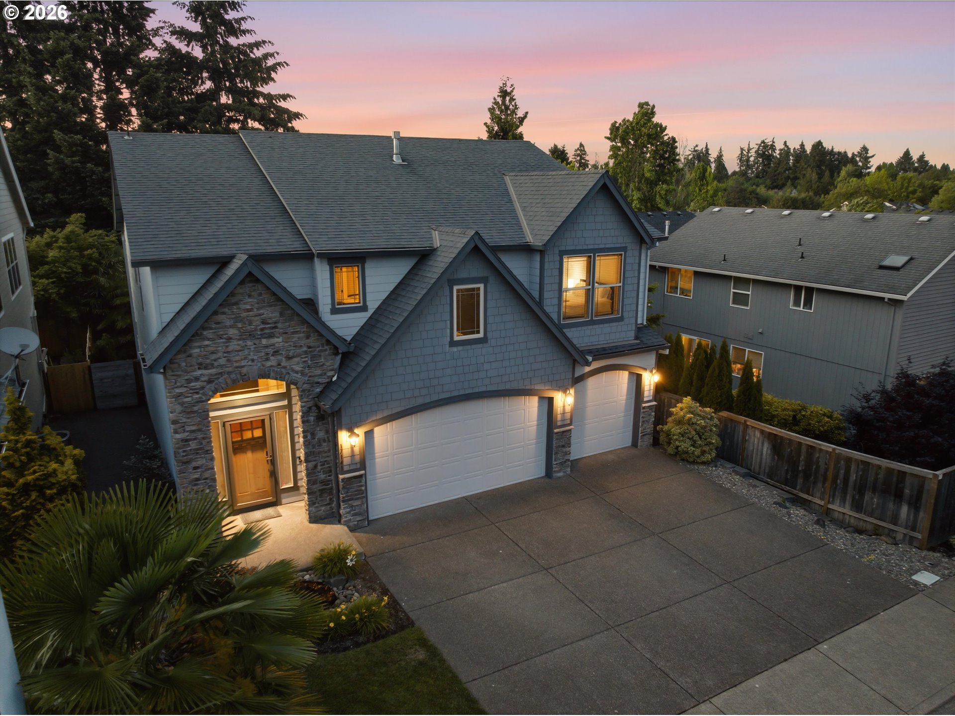 Two-story blue-gray home with stone entry, three-car garage, and warmly lit windows at dusk.