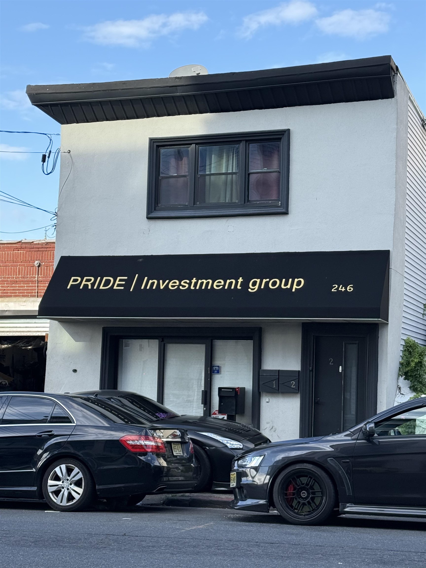 White two-story building with a black PRIDE Investment Group awning and parked cars in front.