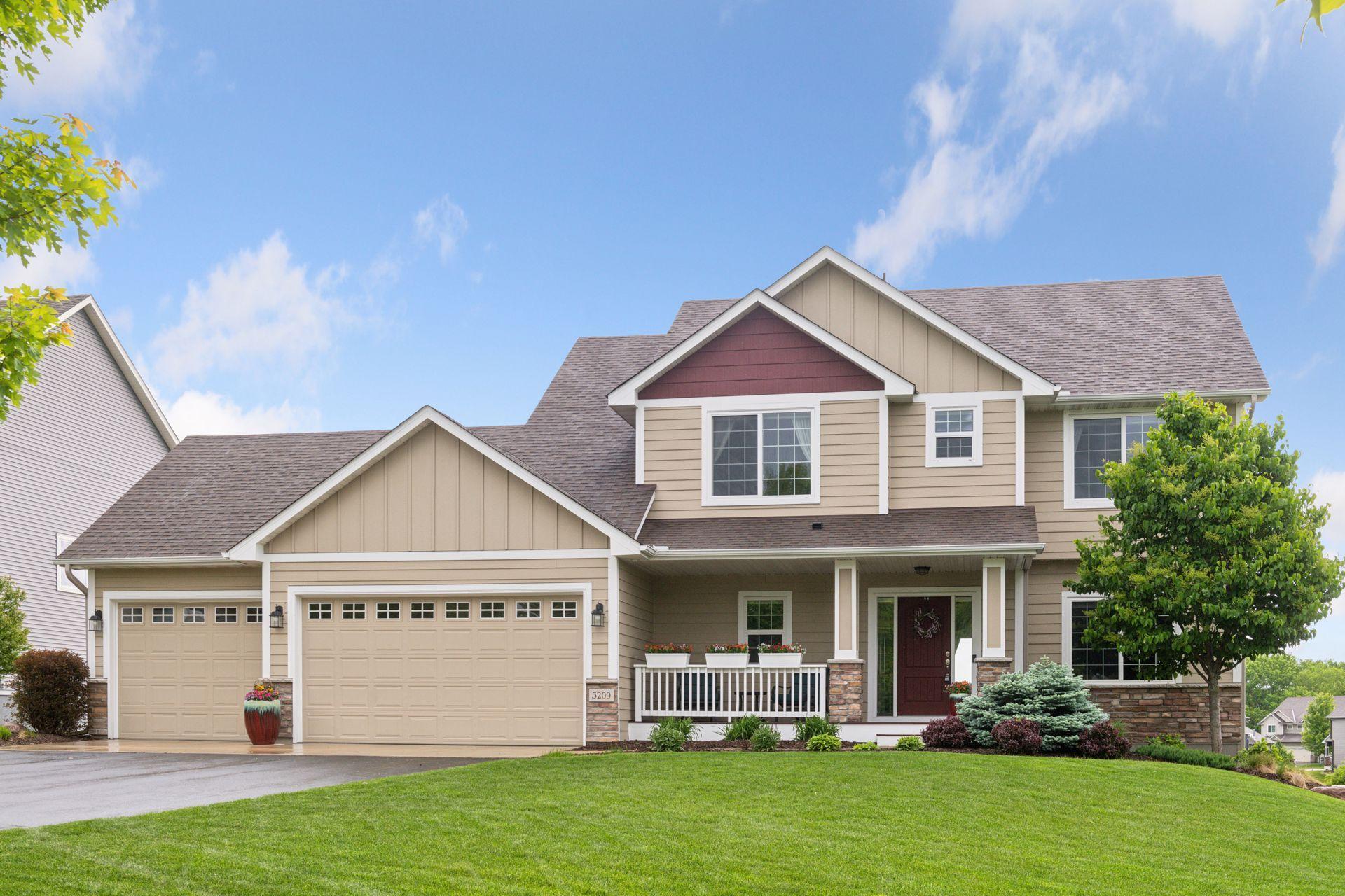 Two-story beige house with a covered porch, three garage doors, and landscaped lawn.