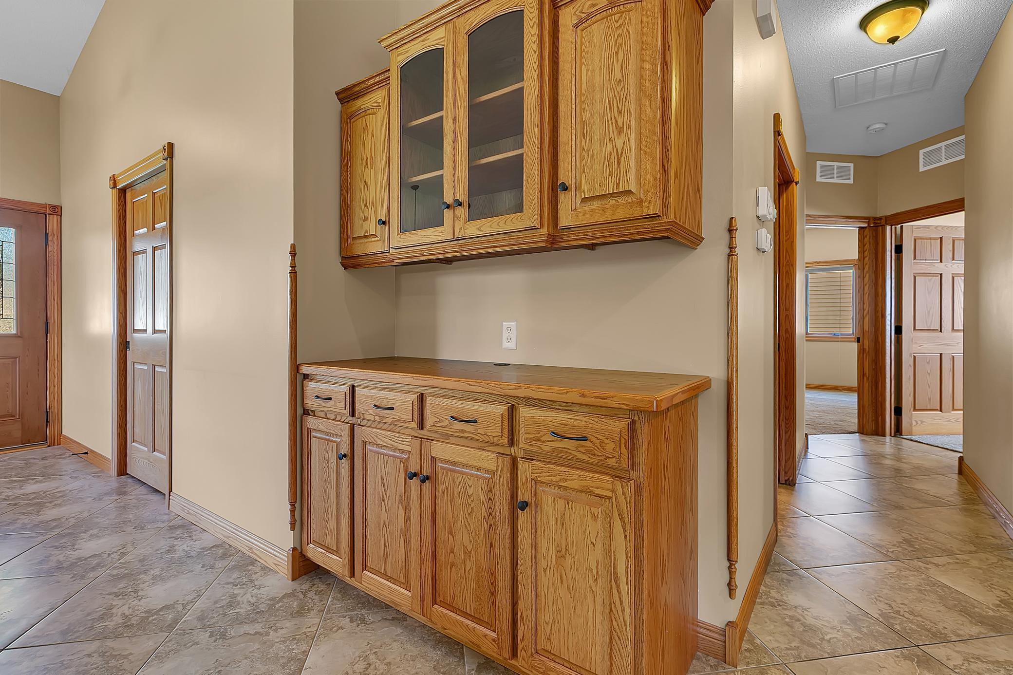 Built-in oak cabinetry and counter beside a tiled hallway with wood-trimmed doorways.