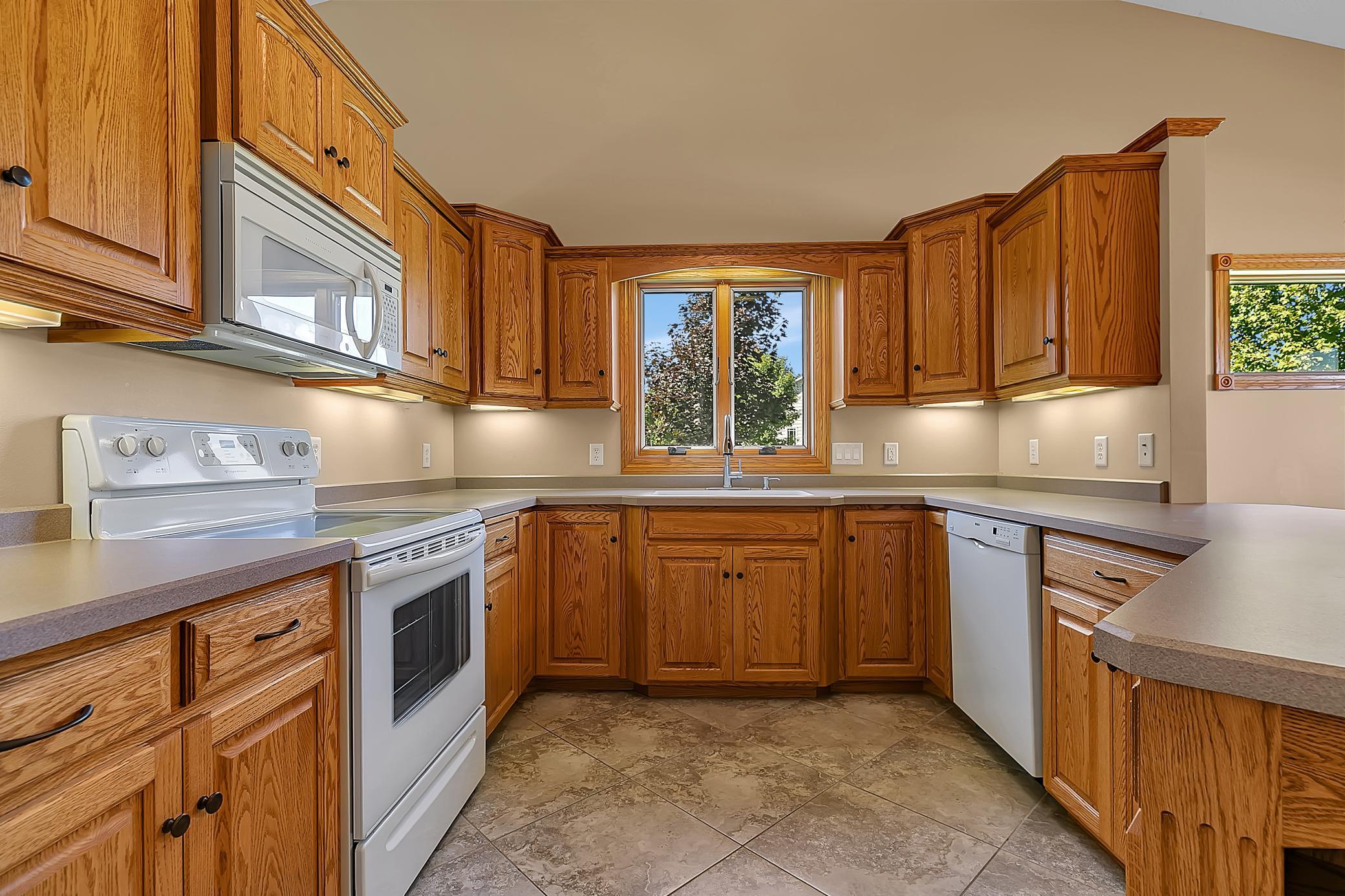 Kitchen with honey-toned wood cabinets, gray counters, tiled floor, and a sink beneath a window.