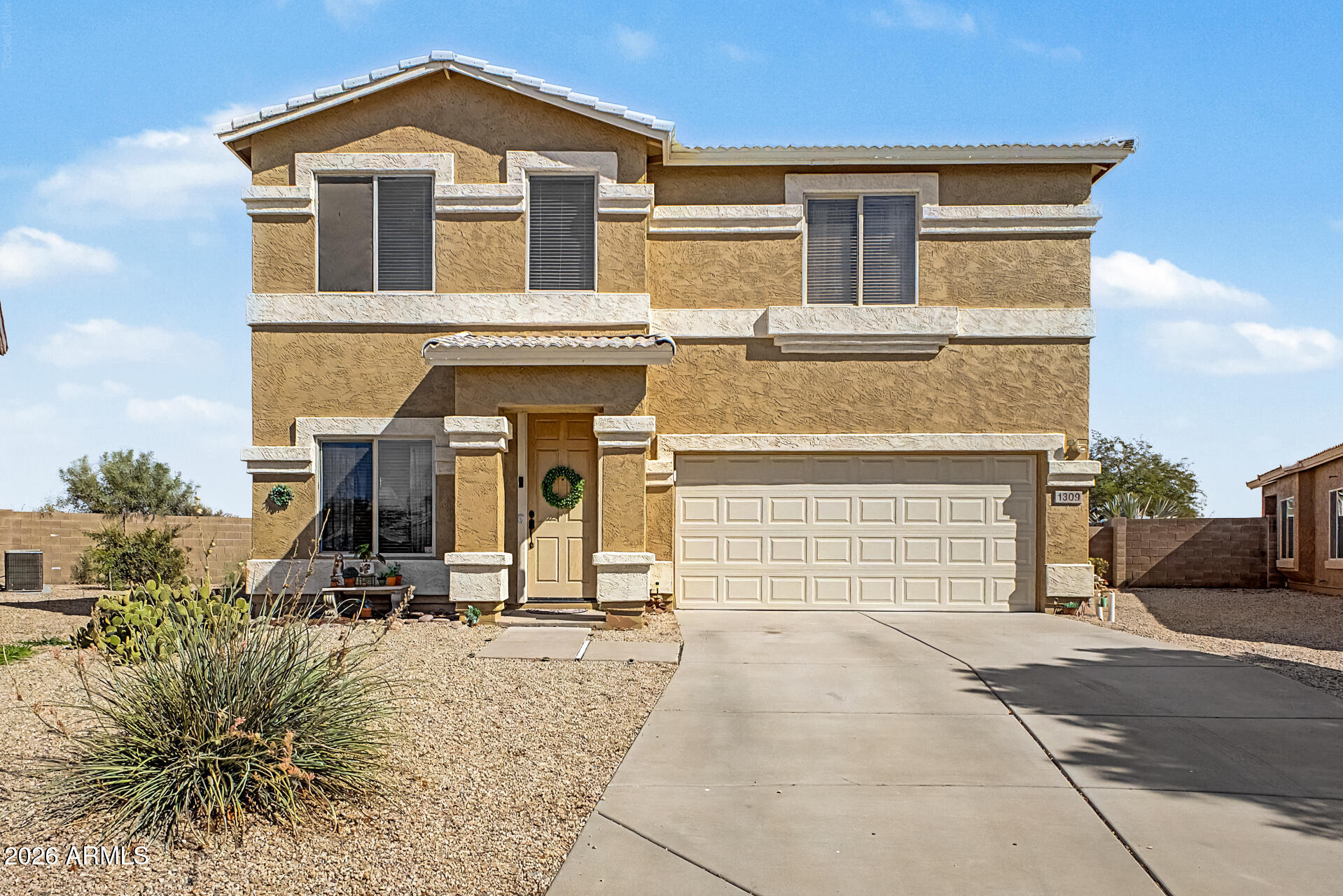 Two-story tan stucco house with stone trim, a double garage, and desert gravel landscaping.