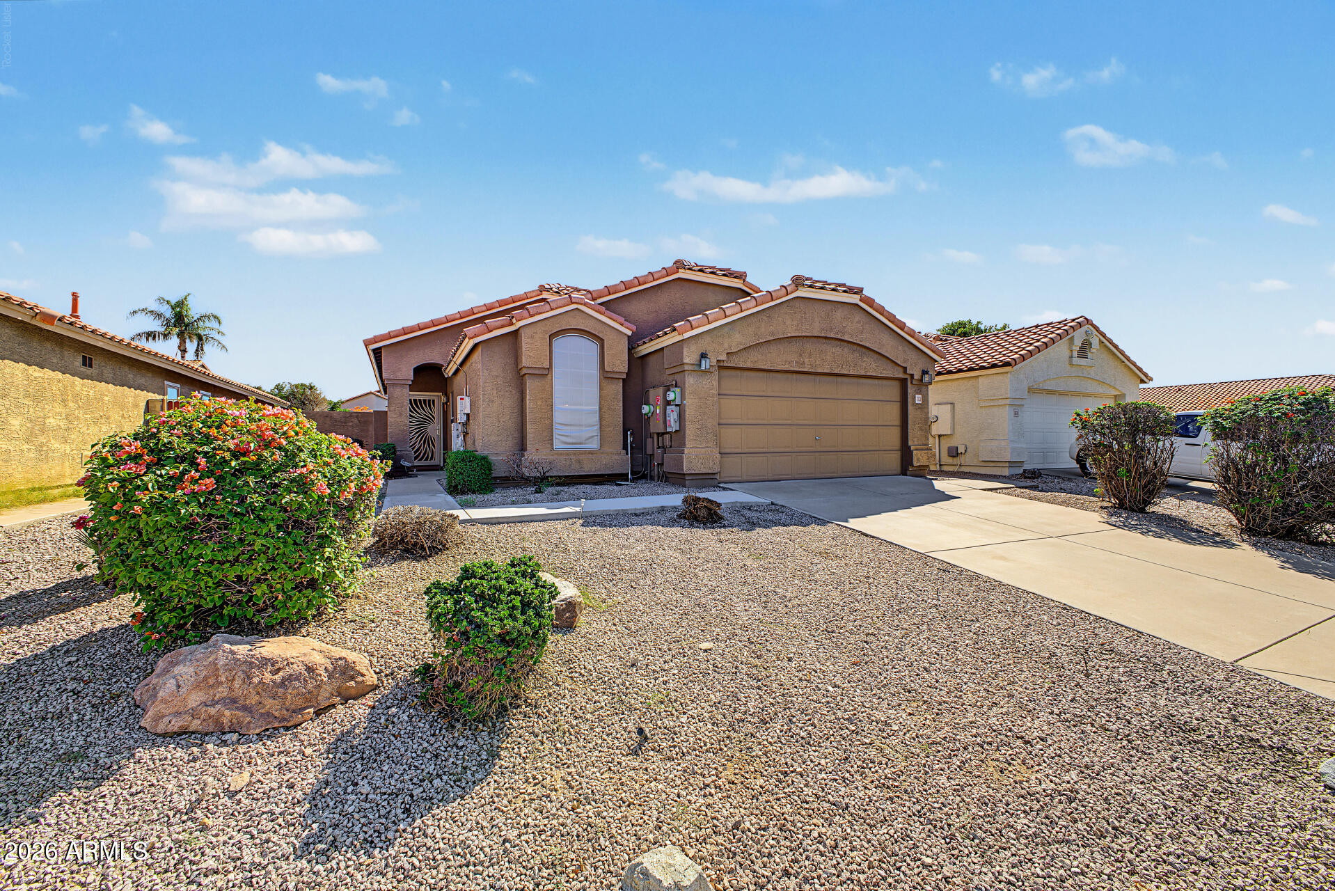 Tan stucco home with a red tile roof, two-car garage, and gravel landscaping.
