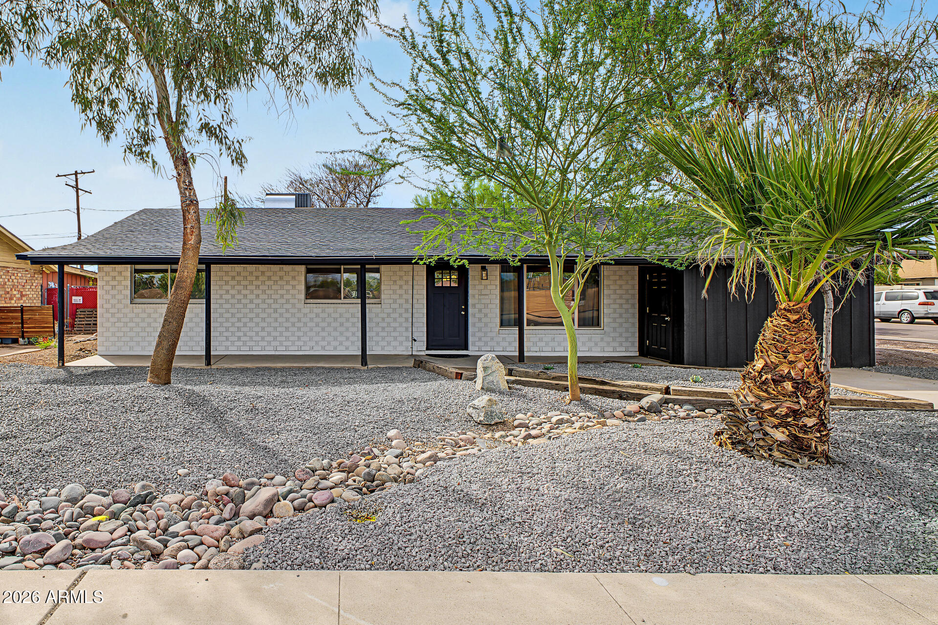 Single-story block home with gravel landscaping, a large palm, and mature desert trees.