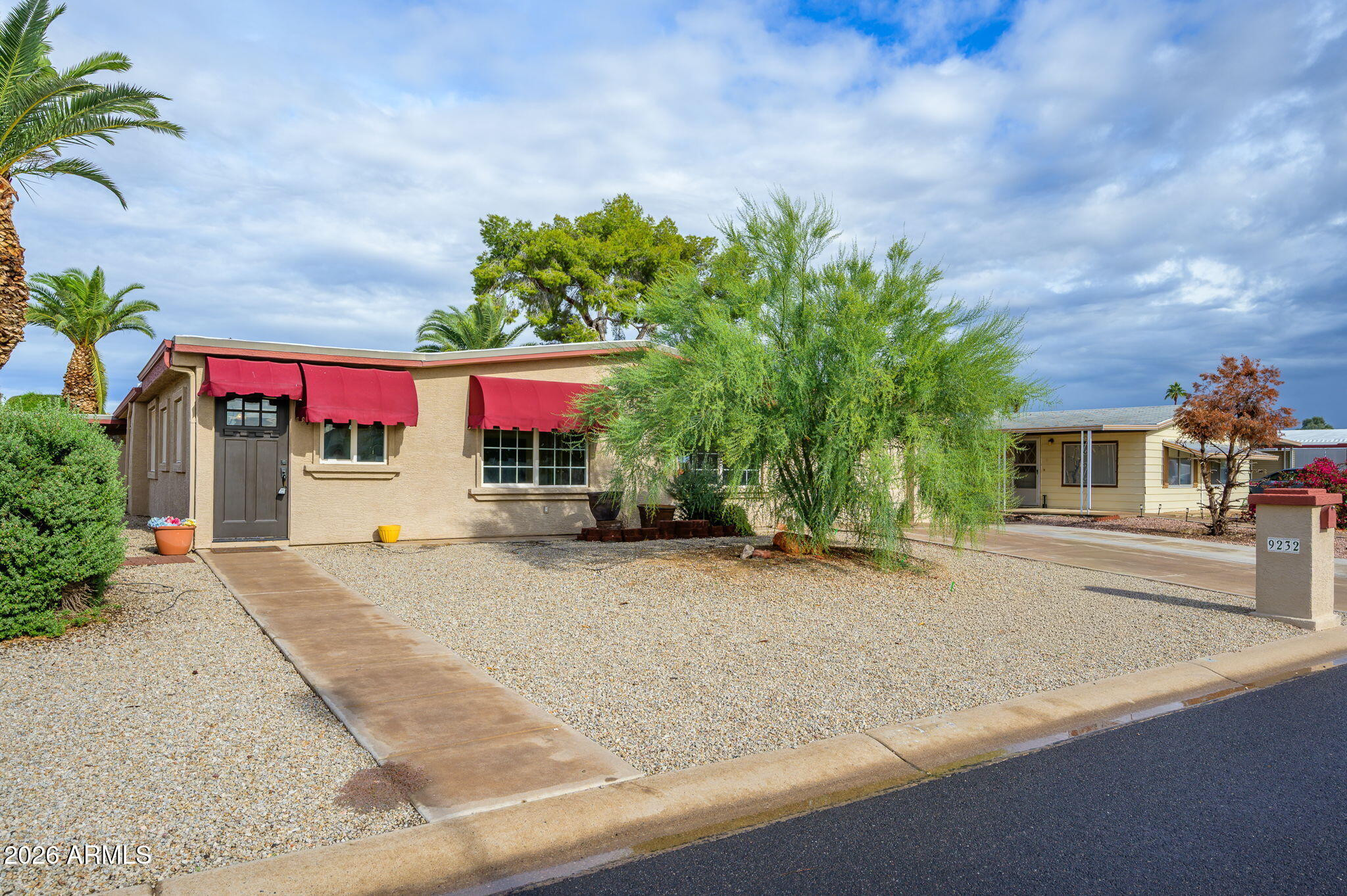 Single-story beige home with red awnings, gravel landscaping, and a concrete walkway.