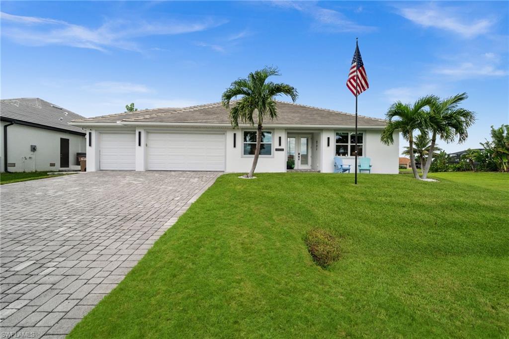 Single-story white home with a paver driveway, palm trees, and an American flag in the front yard.