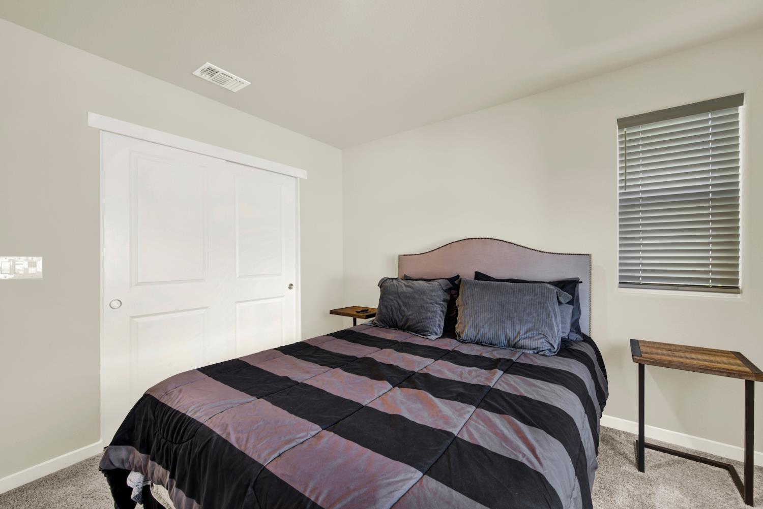 Furnished bedroom with a gray patchwork bedspread, sliding closet doors, and a window with blinds.