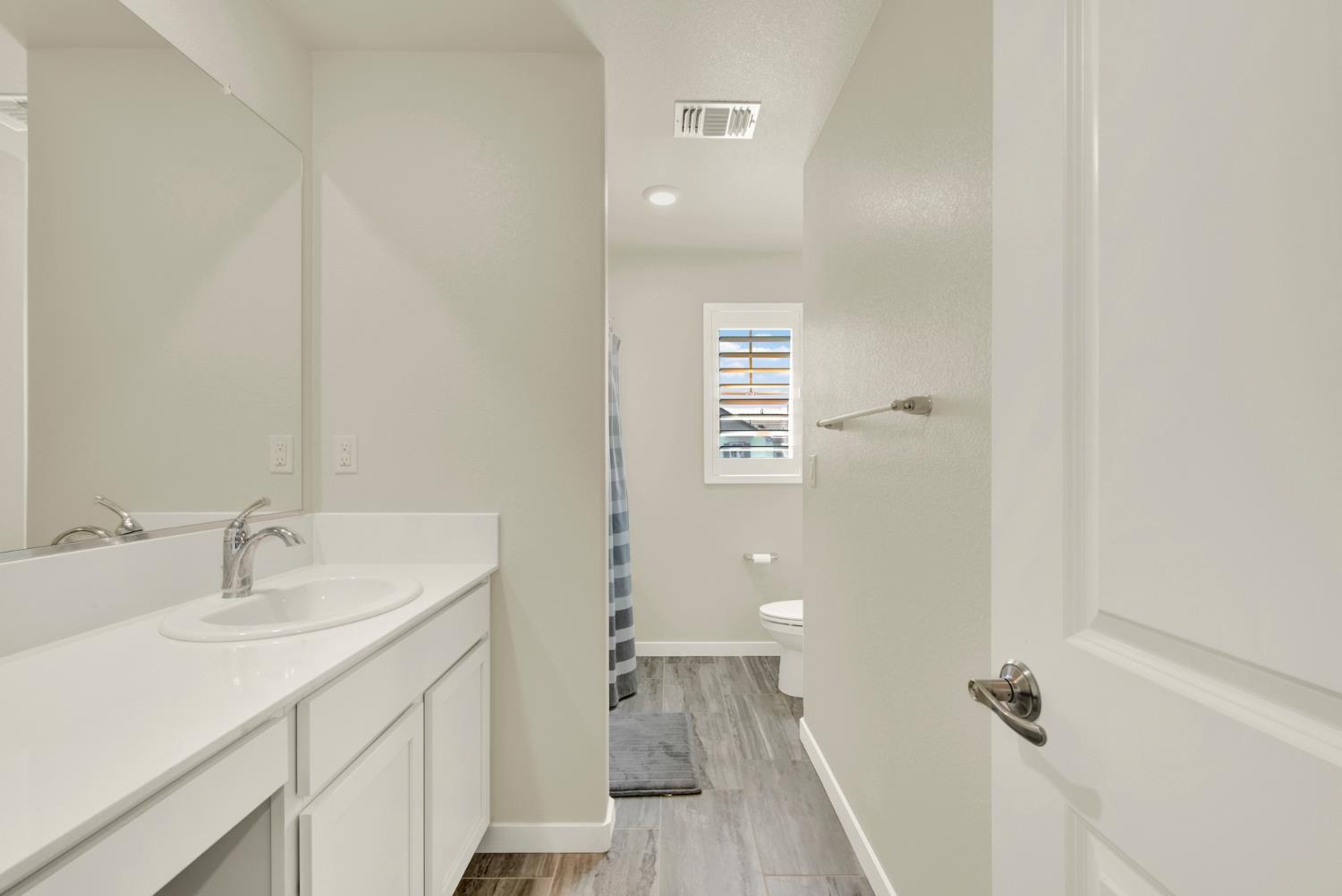 Bathroom with a long white vanity and mirror beside a toilet area with gray tile and a shuttered window.