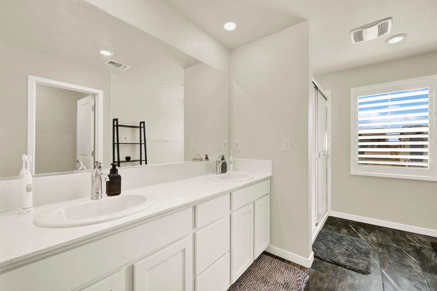 Bright bathroom with a double-sink vanity, large mirror, glass shower, and shuttered window.