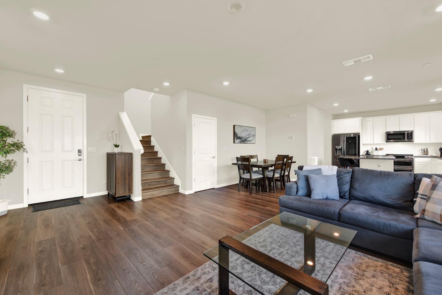 Open-plan living room with a blue sectional, dark wood floors, staircase, dining area, and white kitchen.