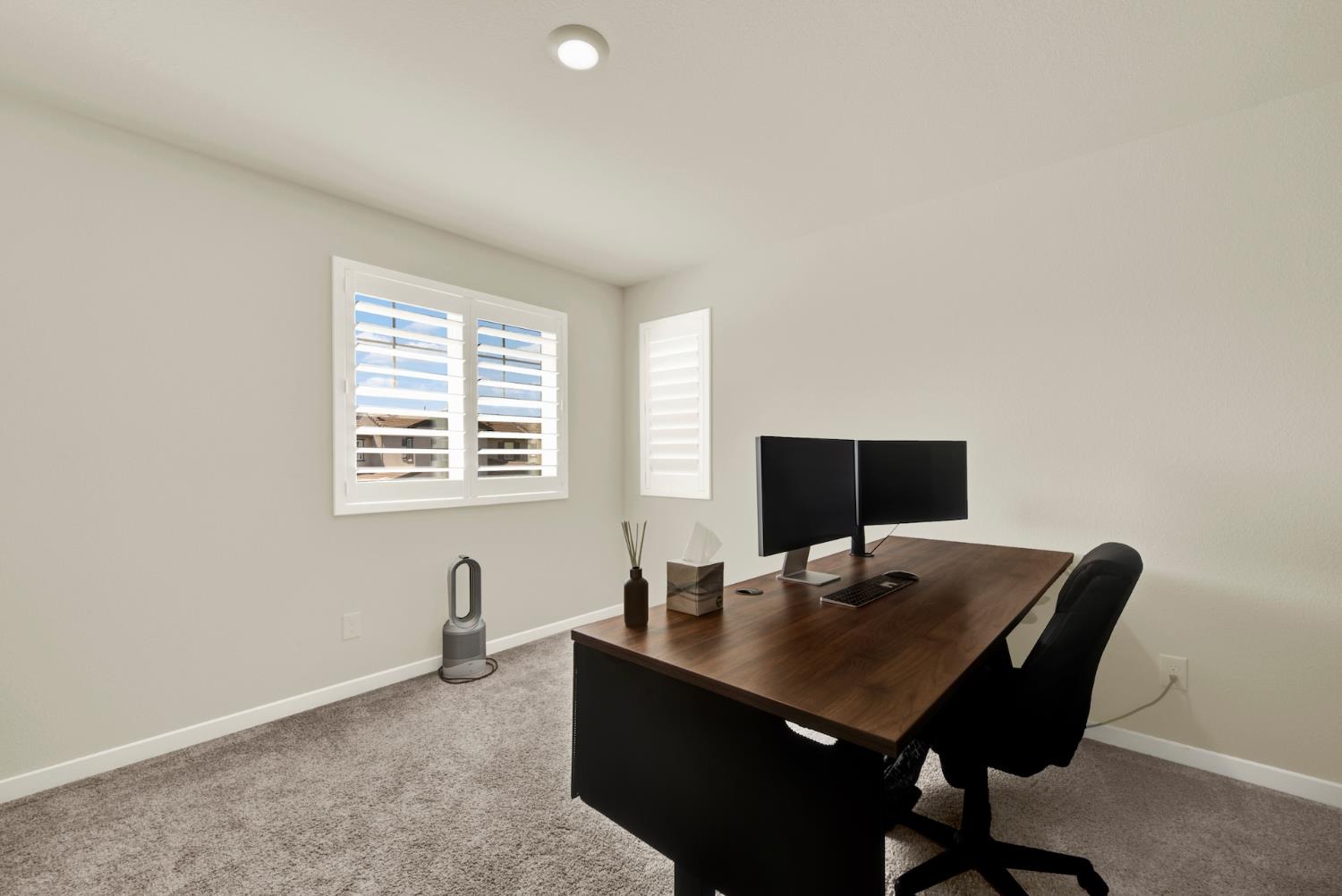 Home office with a large wood desk, dual monitors, and plantation-shuttered windows.