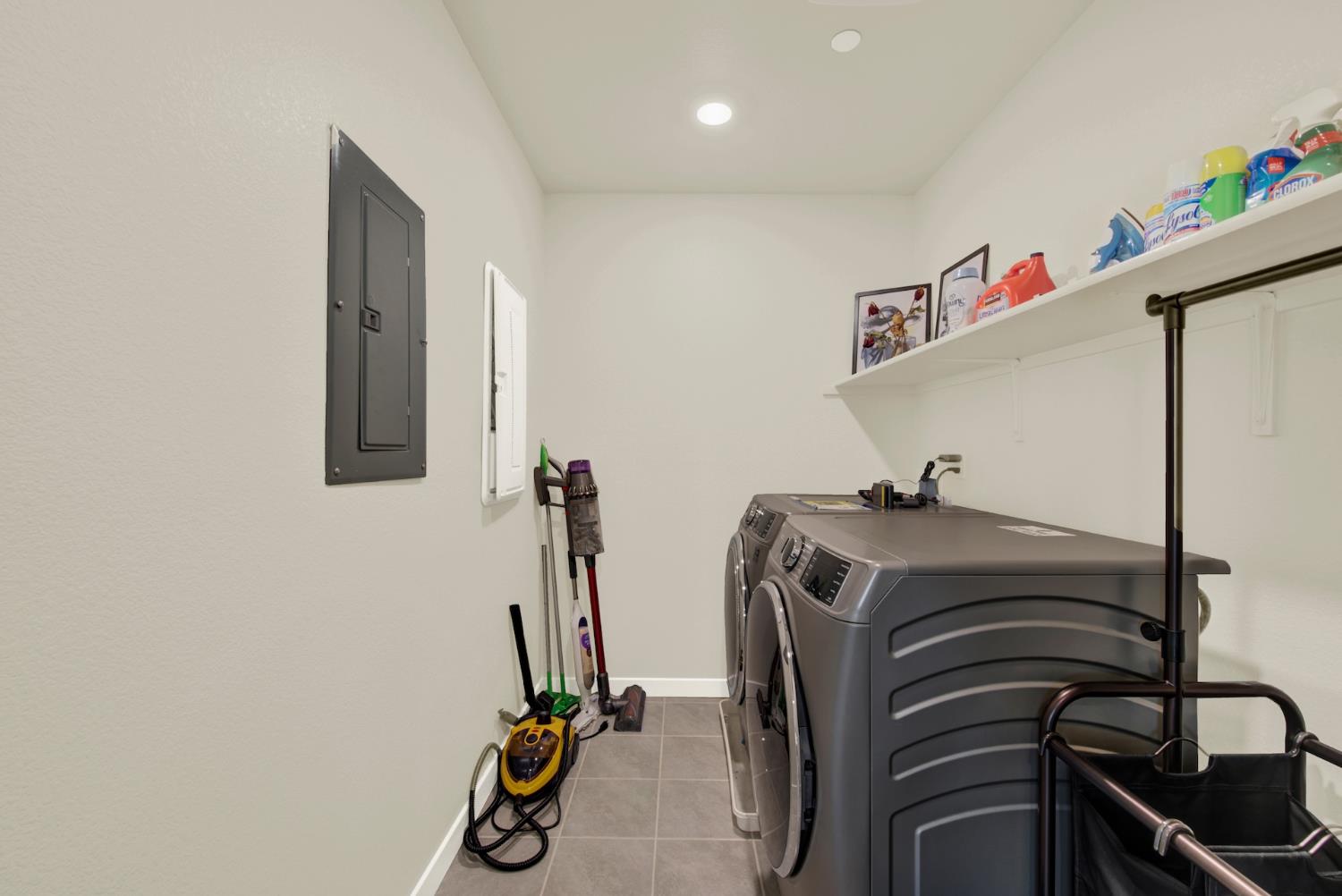 Laundry room with gray front-loading washer and dryer beneath shelves of cleaning supplies.