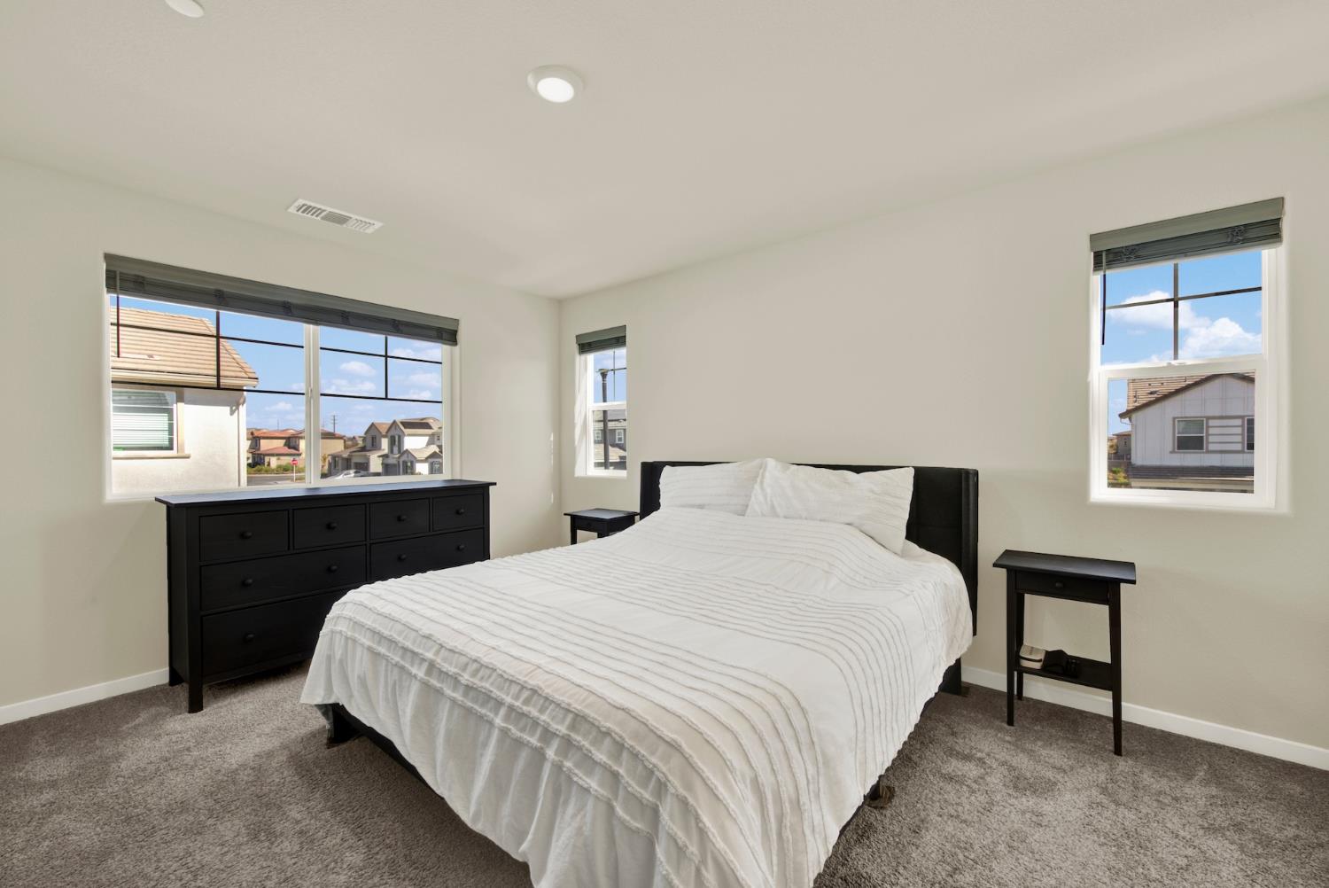 Bedroom with a large bed, white striped bedding, black furniture, and windows overlooking neighboring homes.