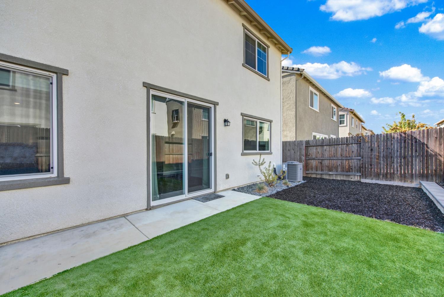 Rear of a two-story home with a sliding door, concrete patio, artificial grass lawn, and fenced yard.