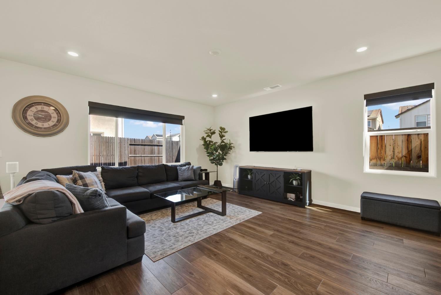 Living room with a dark sectional, wall-mounted TV, wood-look floors, and windows facing a fenced yard.