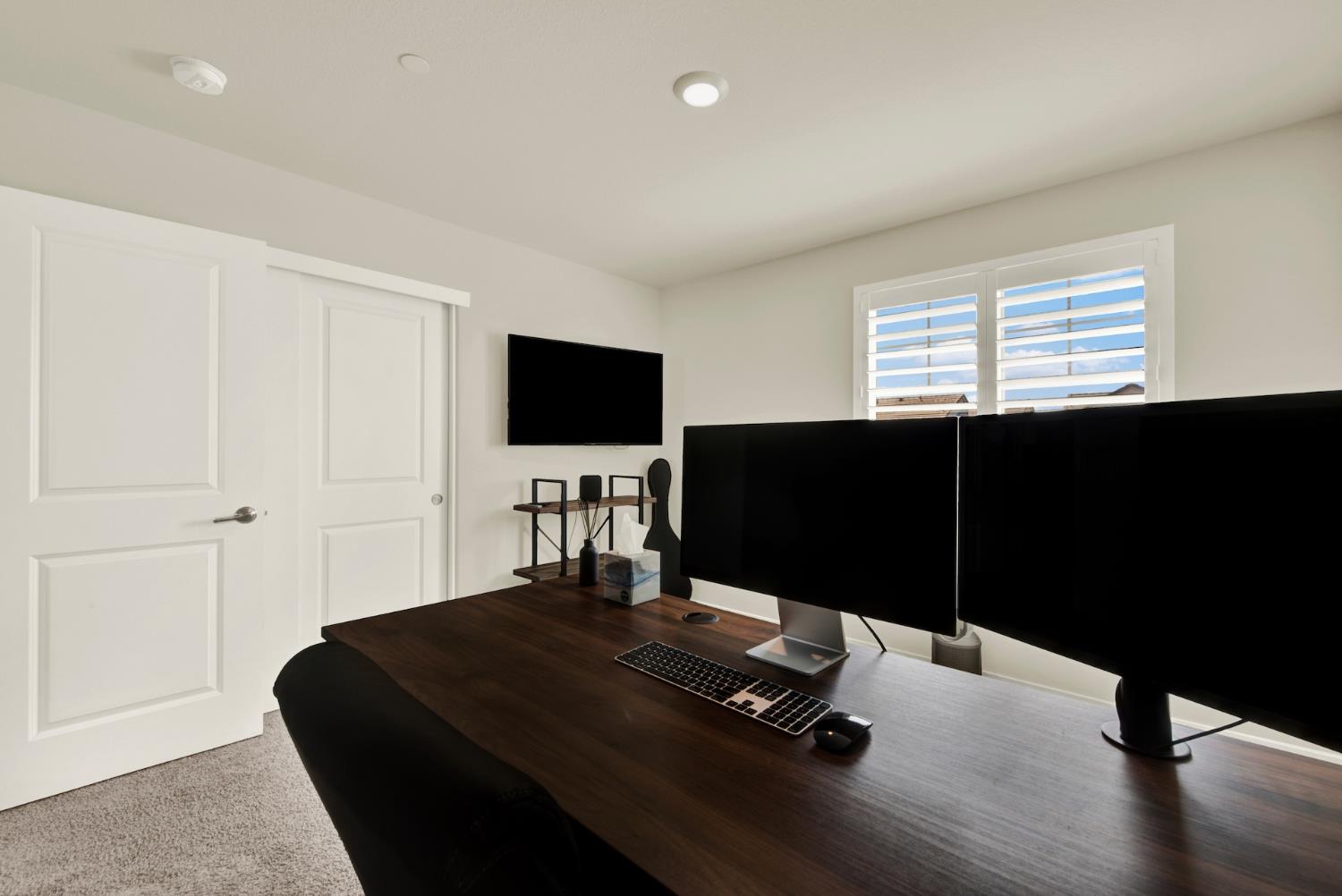 Home office with a dark wood desk, dual monitors, and white plantation shutters.