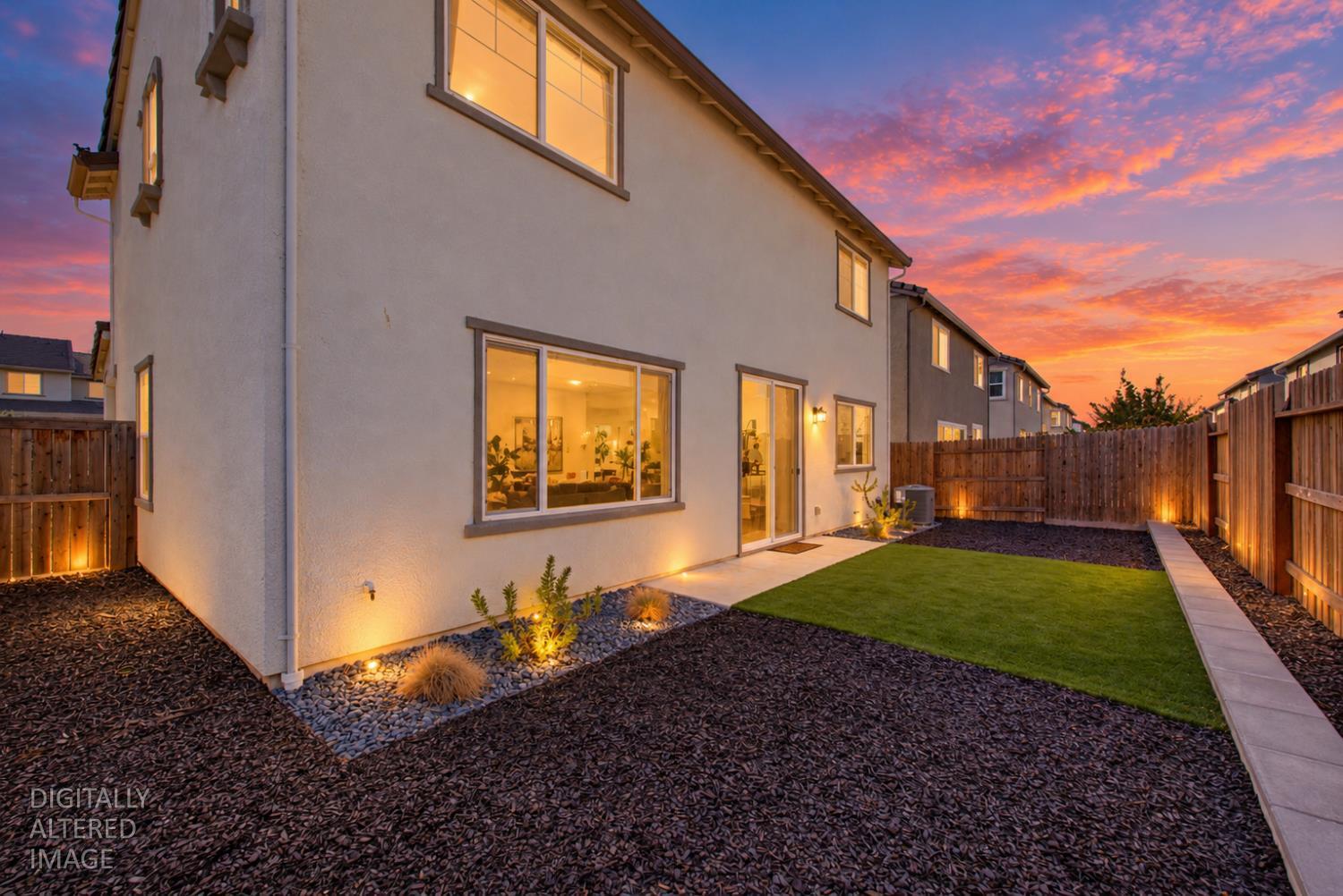 Two-story home’s rear exterior at sunset, with a fenced lawn, landscaped beds, and warmly lit windows.