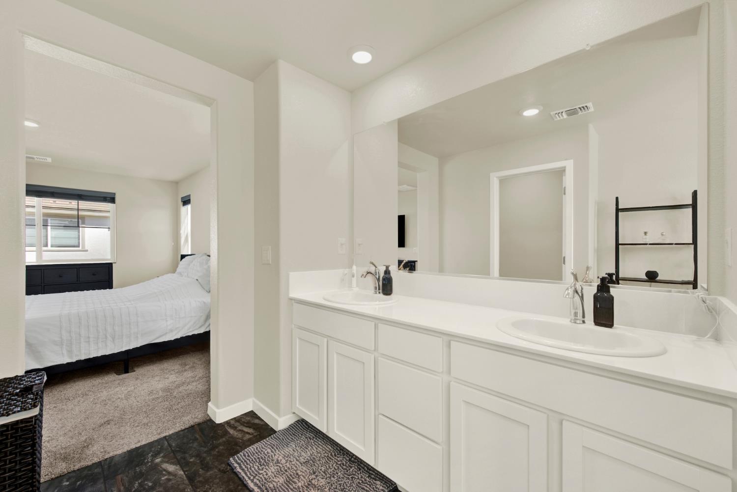 Bathroom with a long double-sink vanity and mirror beside an open doorway to a bedroom.