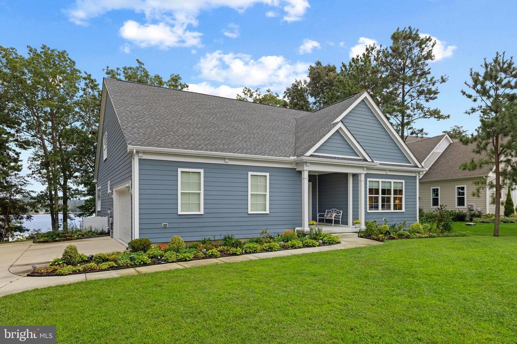 Blue-gray house with a covered porch, landscaped flower beds, broad lawn, and water visible through trees.