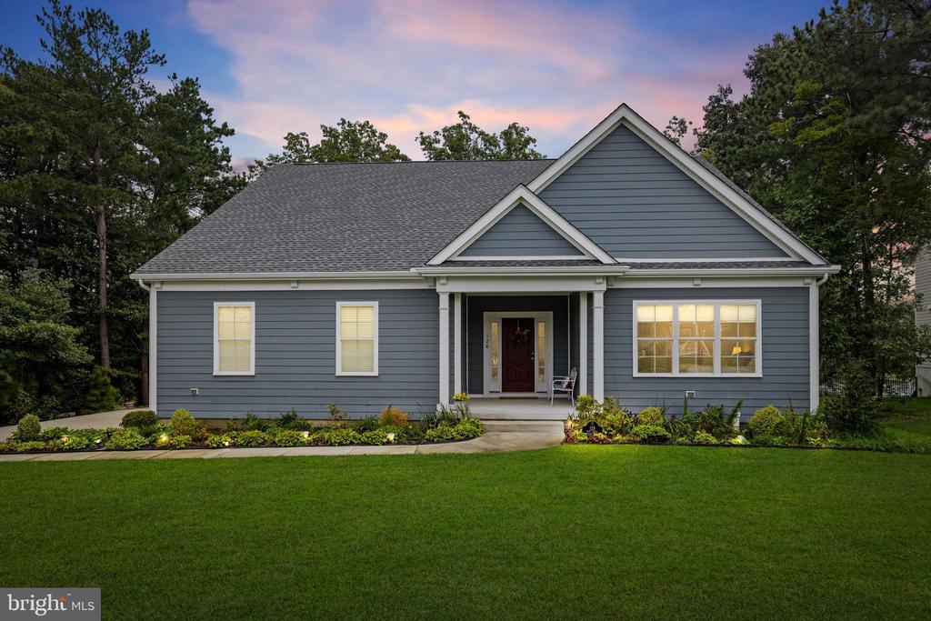 Blue-gray gabled house with a covered entry, glowing windows, landscaped beds, and a broad lawn at dusk.