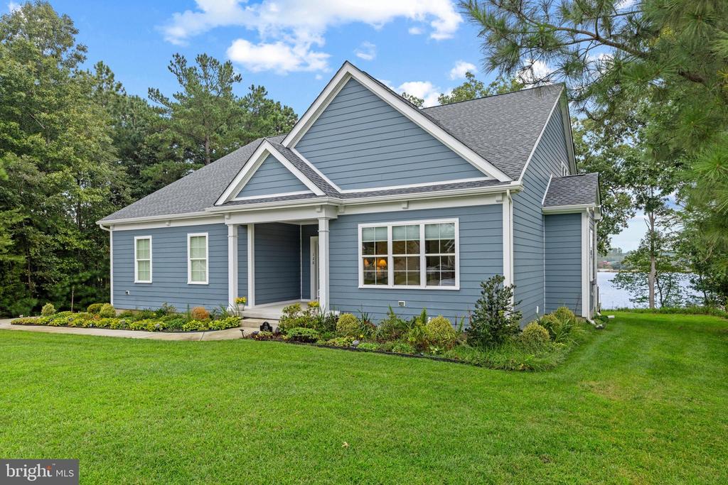 Blue-gray gabled house with white trim, landscaped flower beds, and a glimpse of water beyond the lawn.
