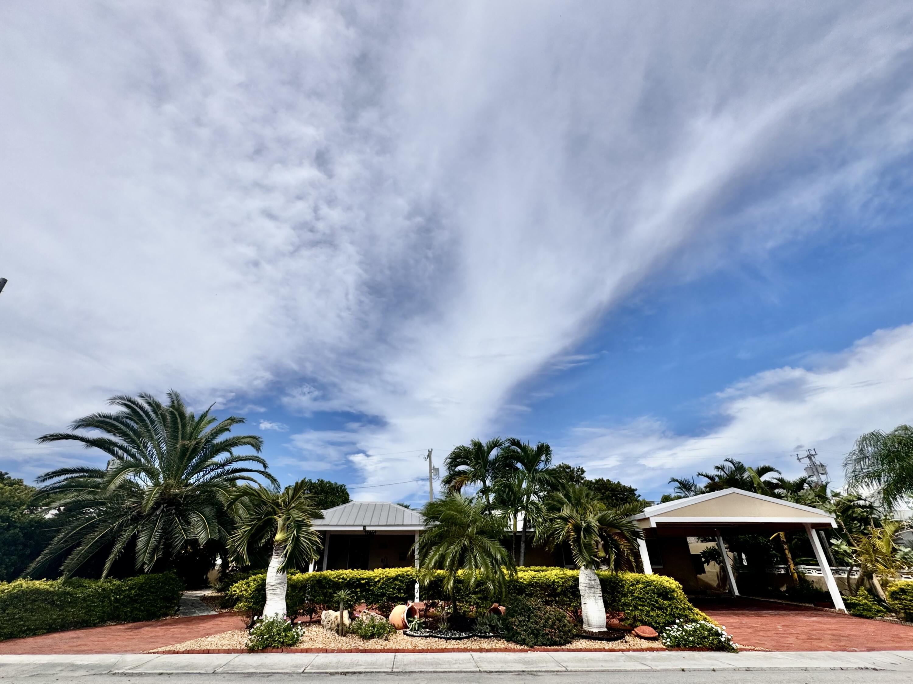 Single-story home with a covered carport, palm trees, clipped hedges, and red-paved driveway.