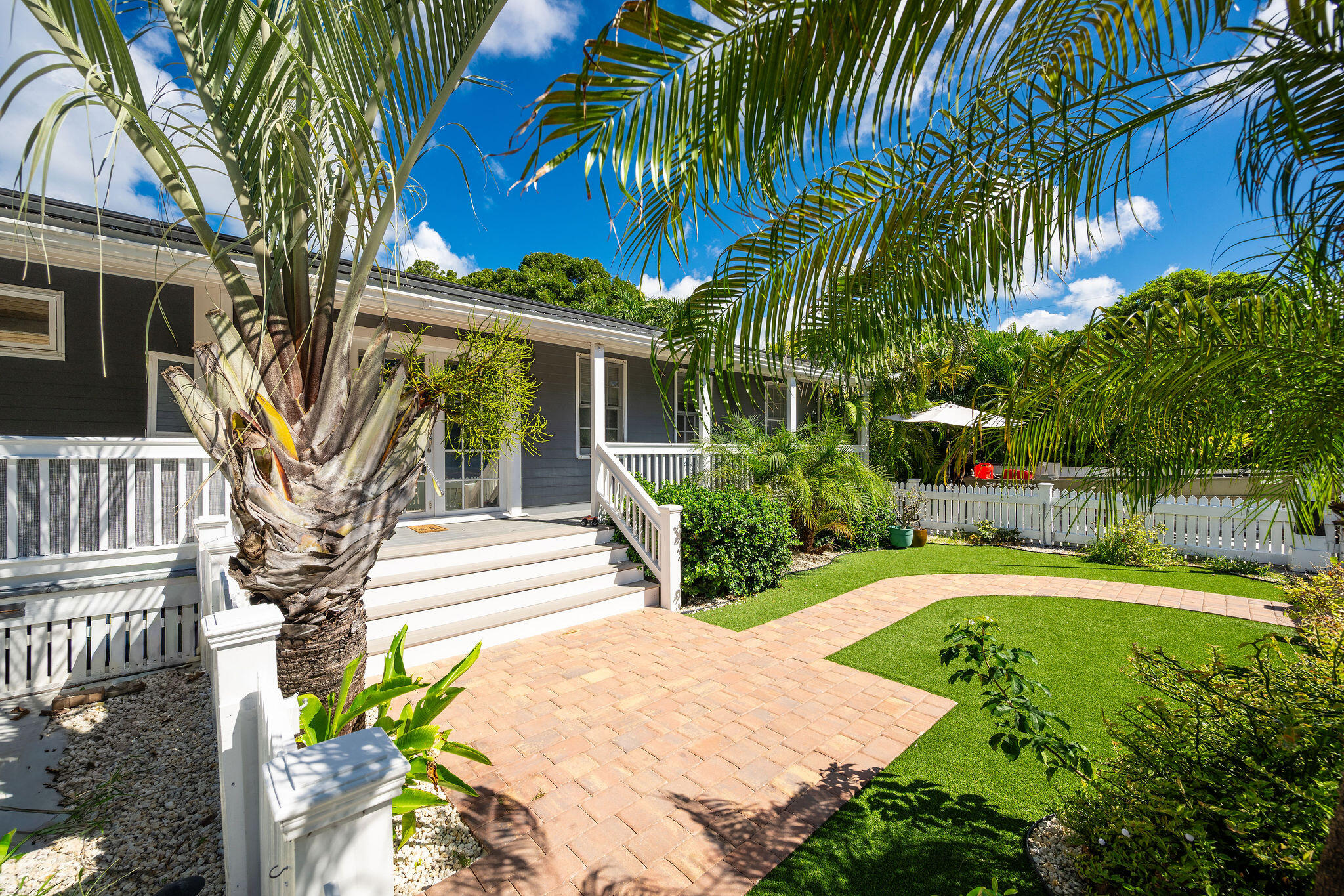 Single-story home with a raised white deck beside a tropical lawn and curving brick path.