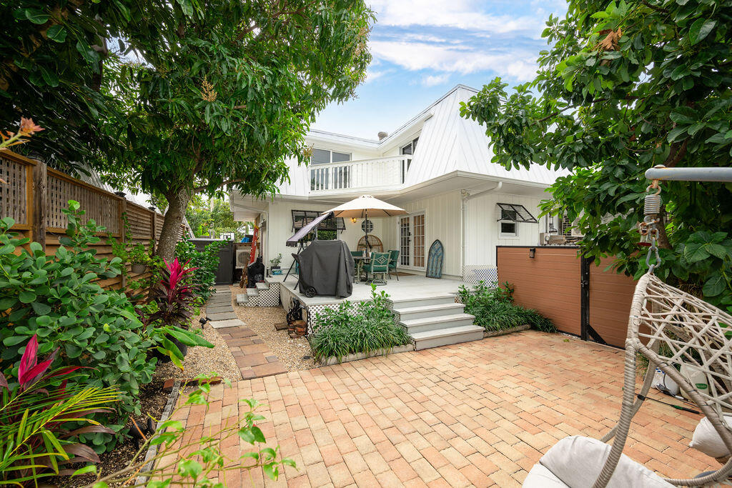 Rear patio of a white two-story home with brick pavers, raised seating area, and tropical landscaping.