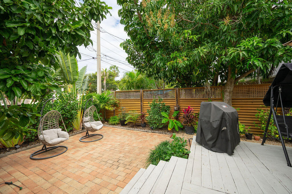 Backyard patio with brick pavers, hanging chairs, tropical landscaping, and a deck with a covered grill.