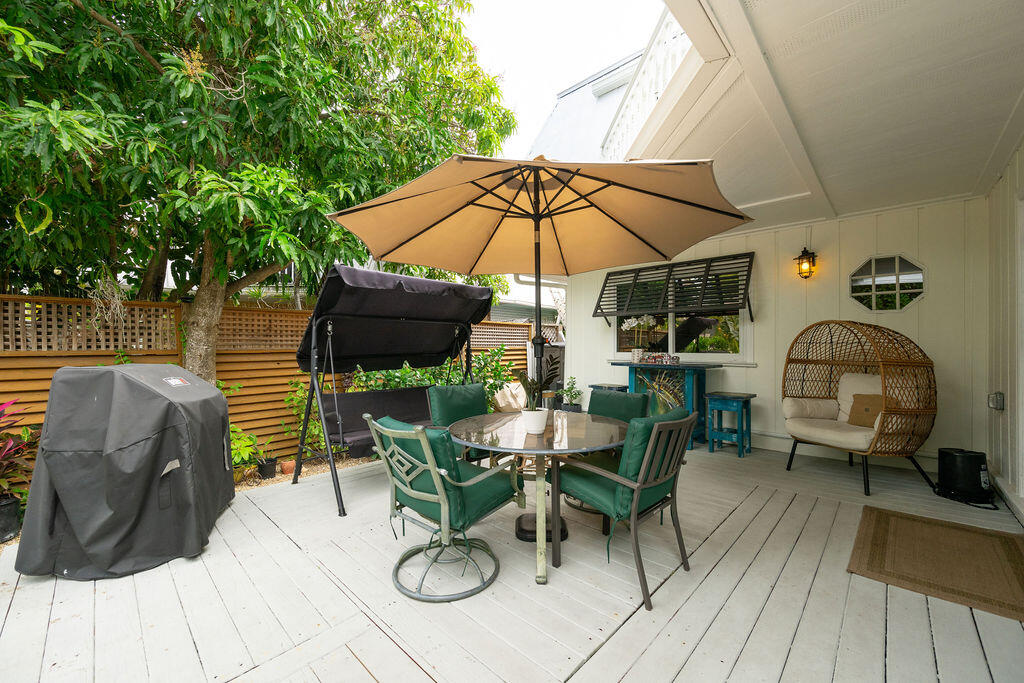 Covered patio with a round dining table, tan umbrella, and hanging swing beside leafy trees.