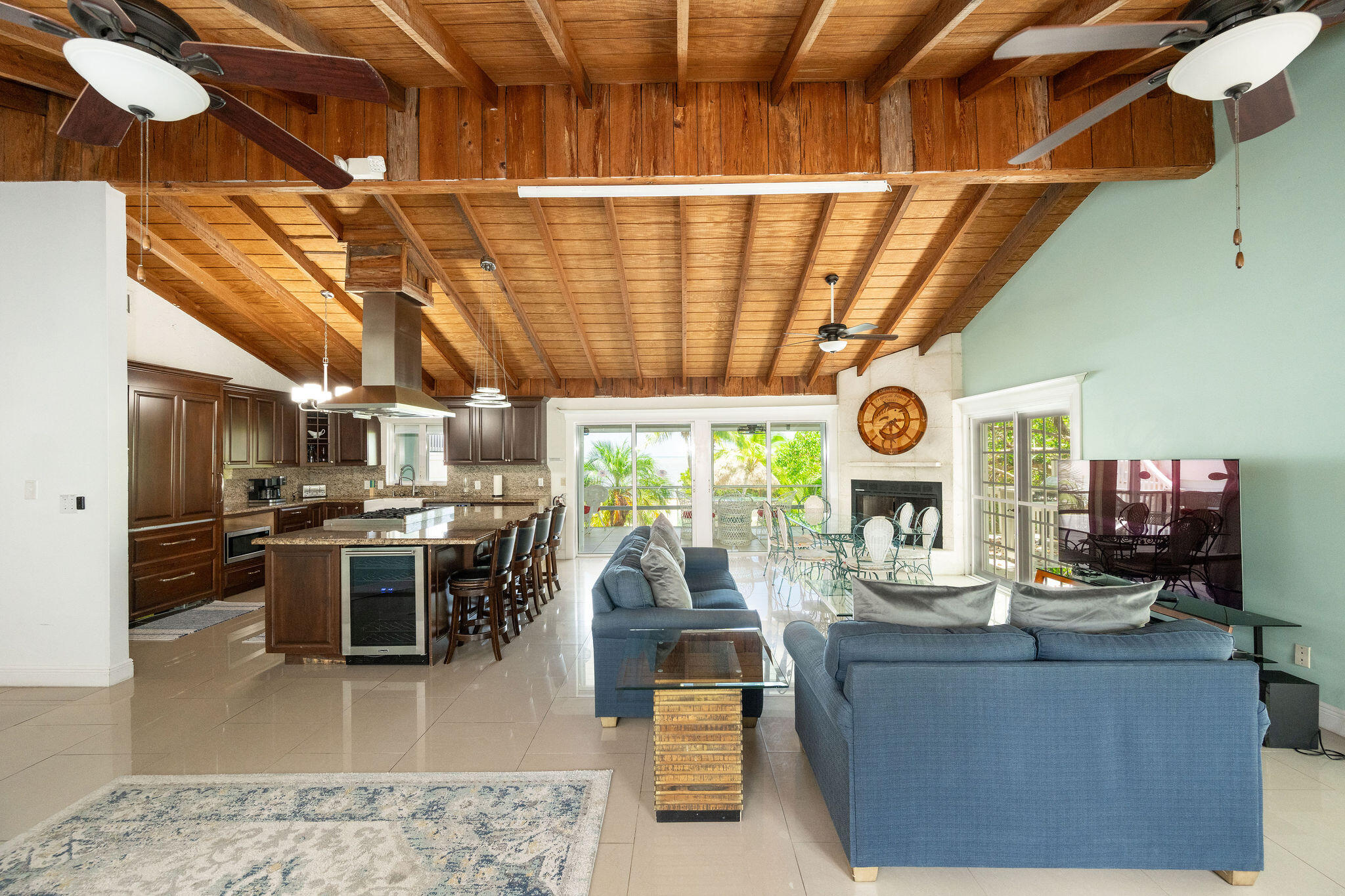Open-plan living room and kitchen beneath a high exposed wood ceiling, with blue sofas and sliding doors to greenery.