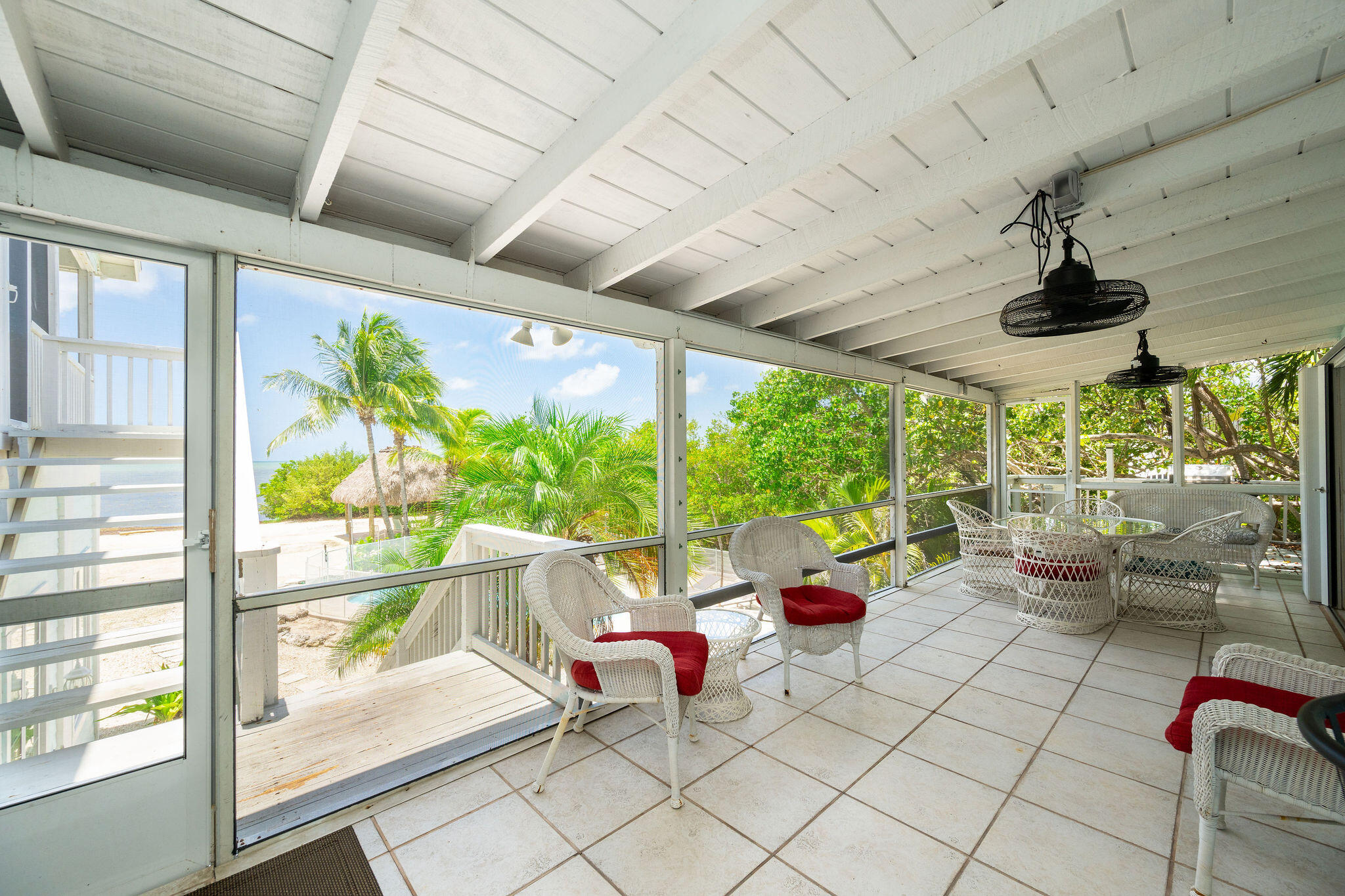 Screened porch with wicker seating and red cushions overlooking palms and water.