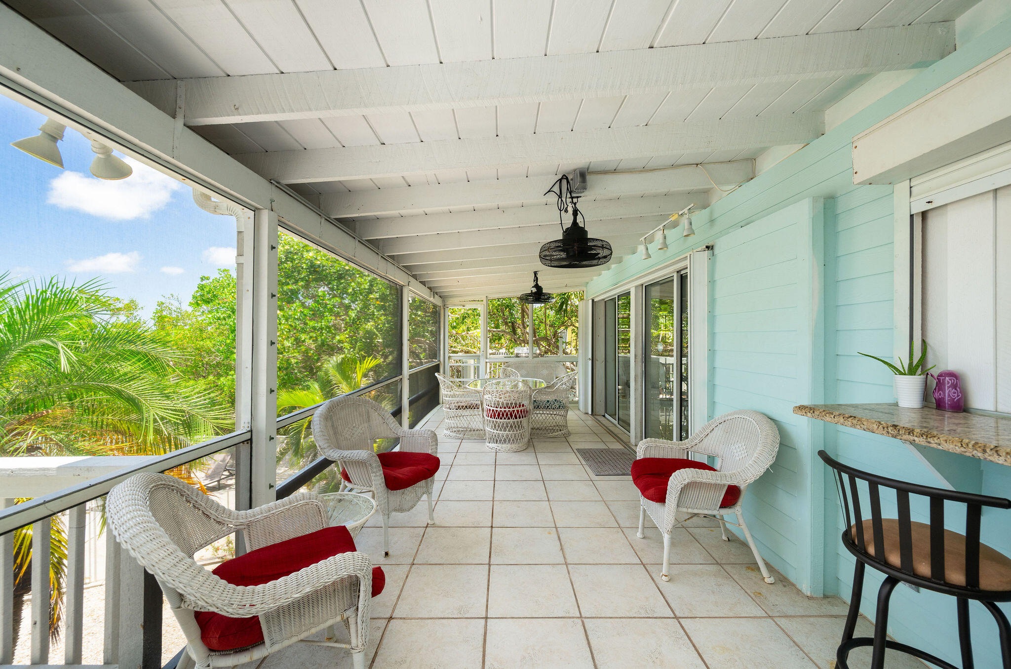 Screened porch with aqua siding, tiled floor, and white wicker chairs with red cushions.