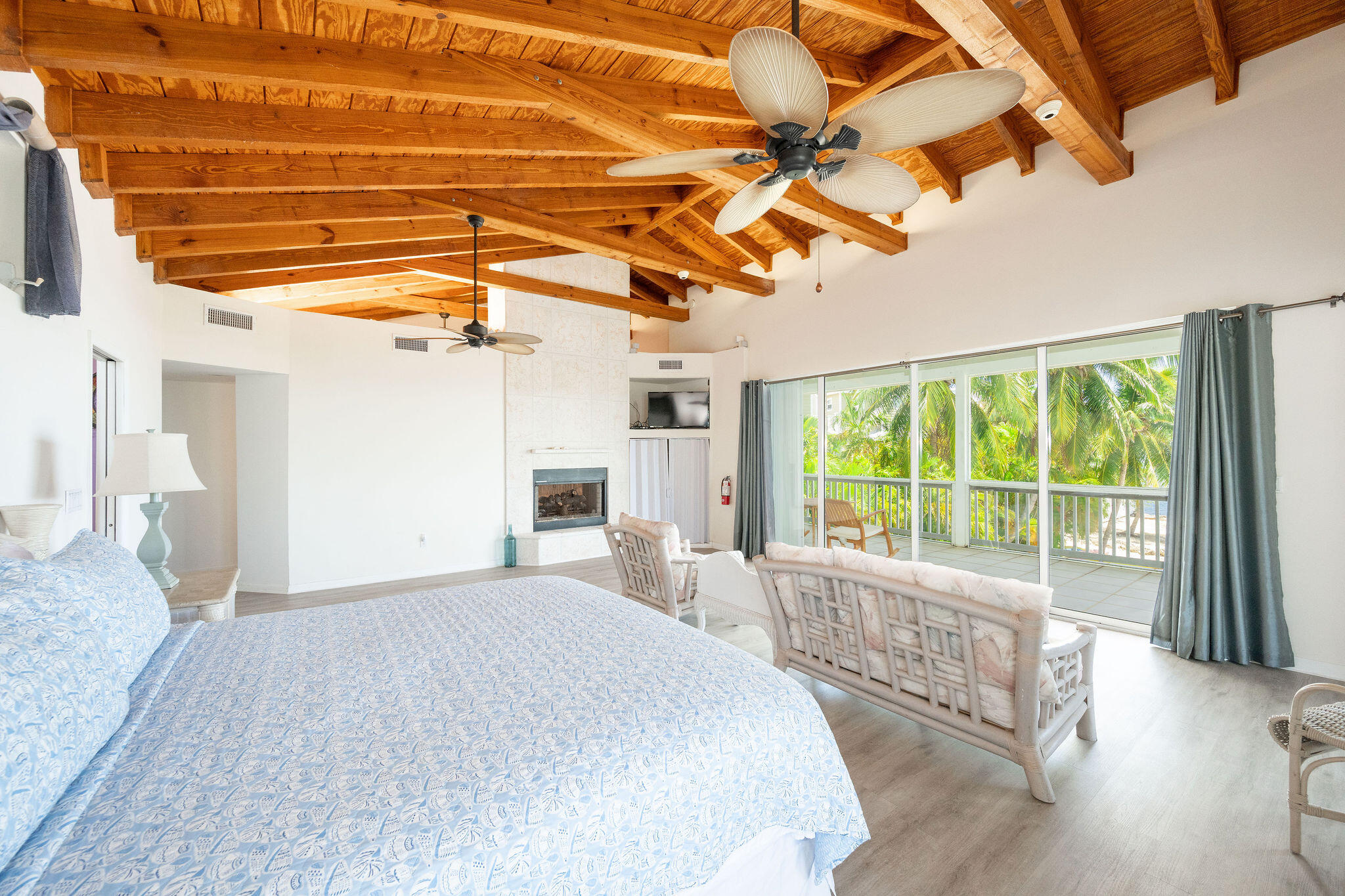 Bedroom with exposed wood ceiling, fireplace, and sliding doors to a palm-lined balcony.