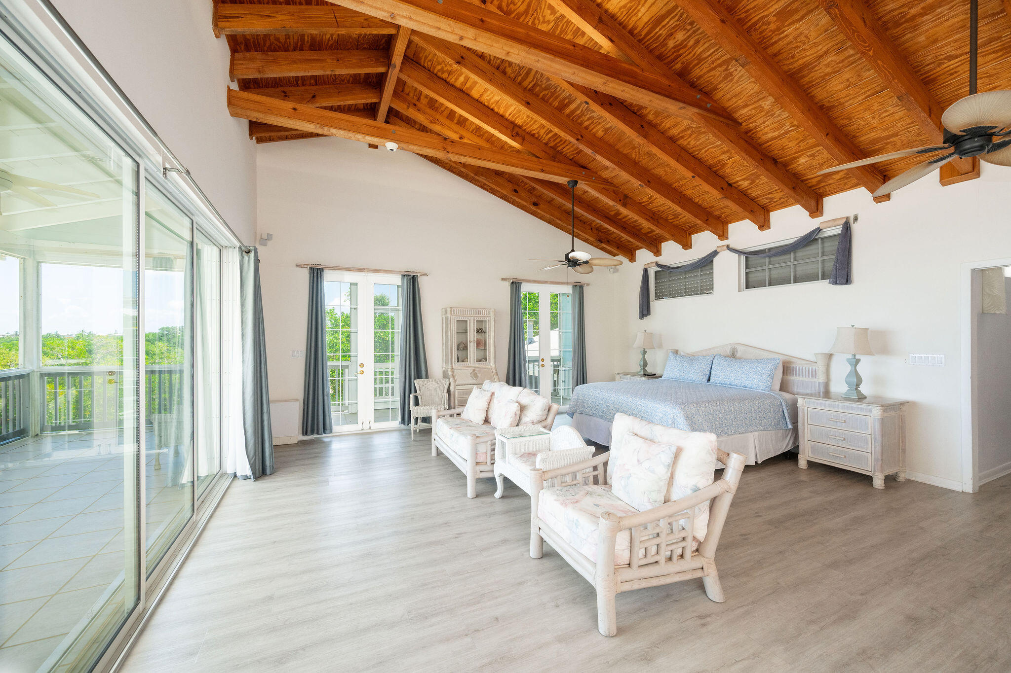 Furnished bedroom with a vaulted wood-beam ceiling, king bed, wicker seating, and sliding doors to a balcony.