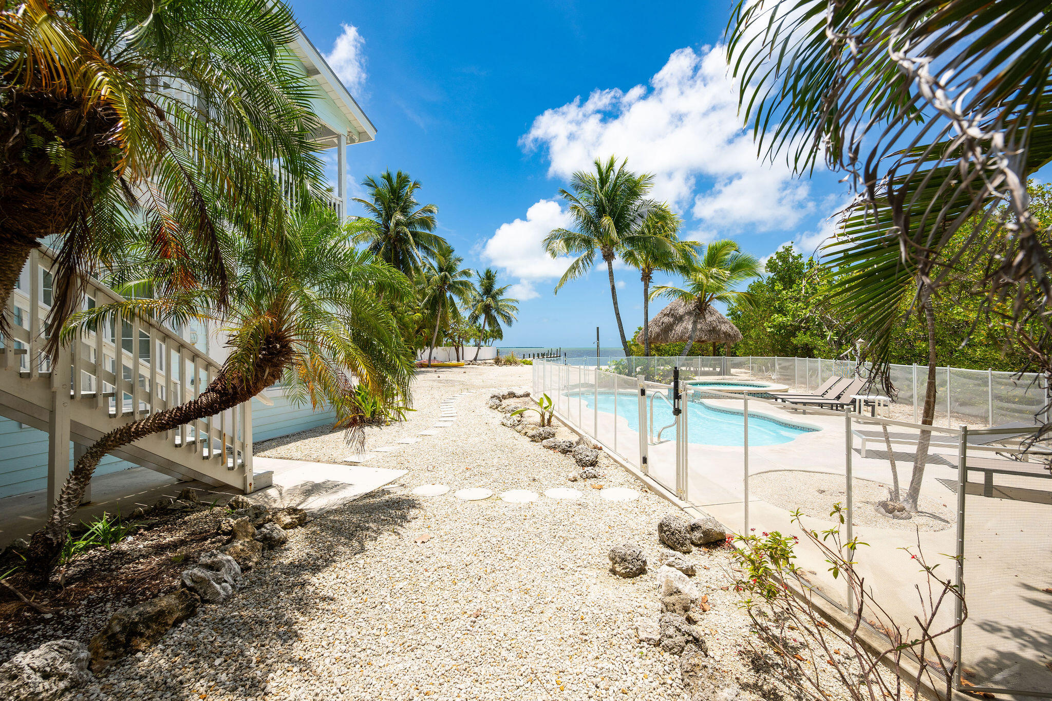 Fenced swimming pool beside a tropical gravel yard, with palms and water visible beyond.