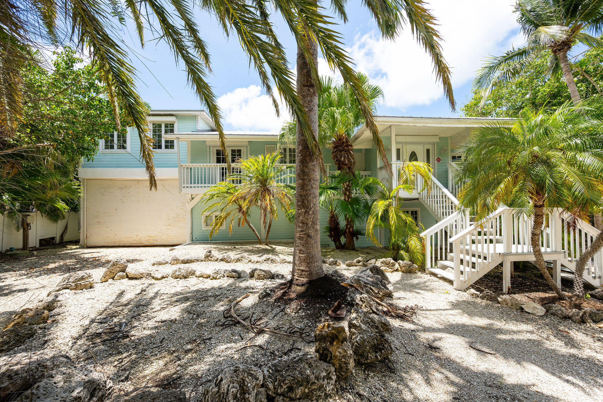 Pale blue raised house with white exterior stairs, framed by palms and rocky landscaping.