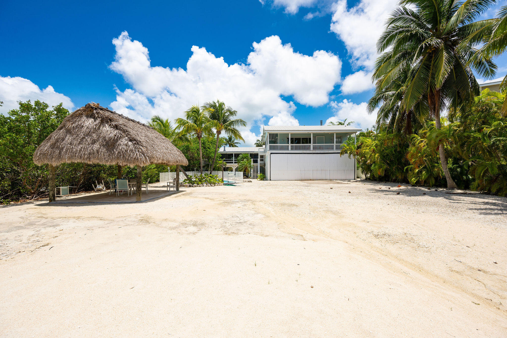 Raised coastal home with a thatched tiki hut, sandy yard, palms, and fenced pool area.