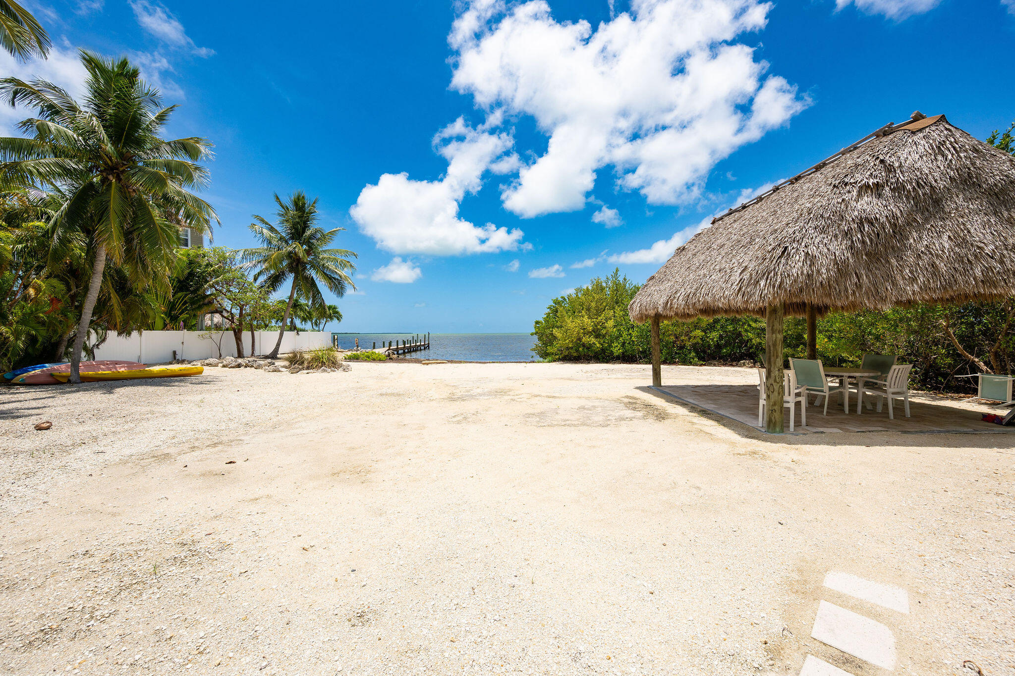 Sandy waterfront yard with a thatched tiki pavilion, palm trees, kayaks, and a dock