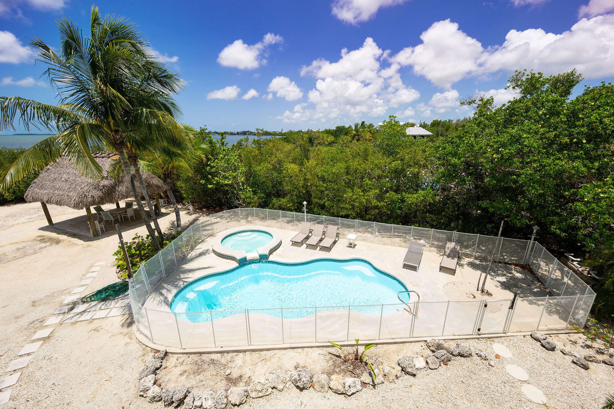Fenced pool and circular spa beside a thatched tiki hut, palms, and waterfront.